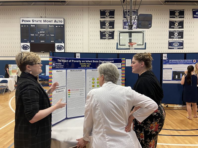 A student talks to others in front of a tri-fold board that reads "The Impact of Poverty on Infant Brain Development"
