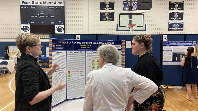 A student talks to others in front of a tri-fold board that reads "The Impact of Poverty on Infant Brain Development"