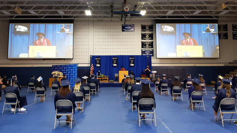 Students in regalia in the campus gym listening to remarks from the chancellor