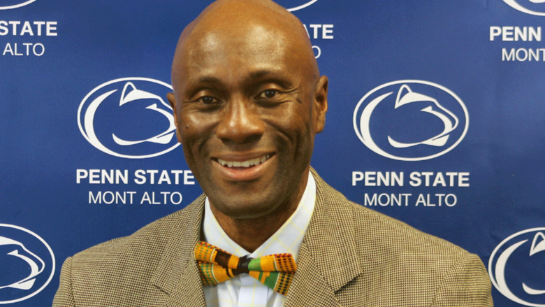 Photo of Francis Achampong in a tie and jacket in front of a blue Penn State backdrop. 