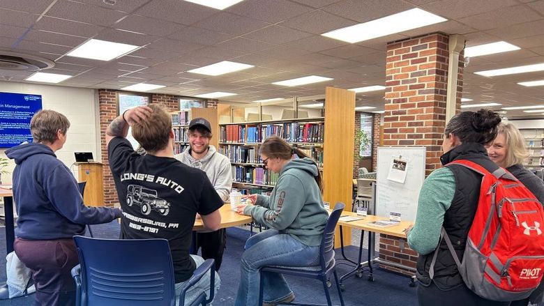 Four people sitting around a table talking and laughing. Two additional people standing to the right talking and laughing.