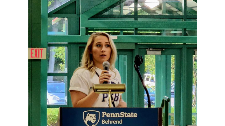 A person stands at a podium giving a speech, the podium has a banner that reads Penn State Behrend