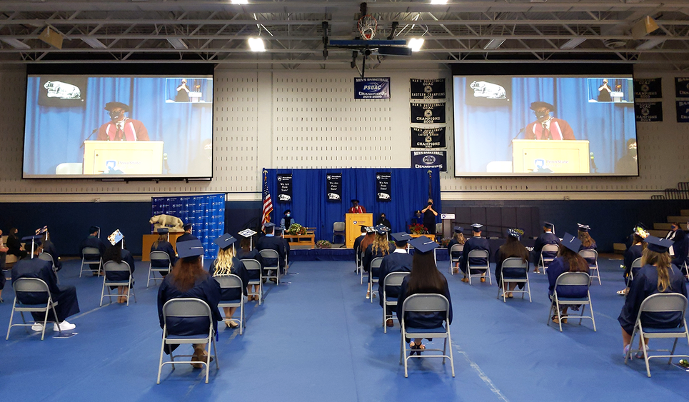 Students in regalia in the campus gym listening to remarks from the chancellor