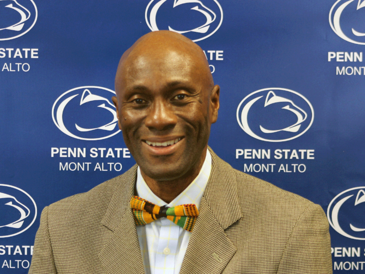Photo of Francis Achampong in a tie and jacket in front of a blue Penn State backdrop. 