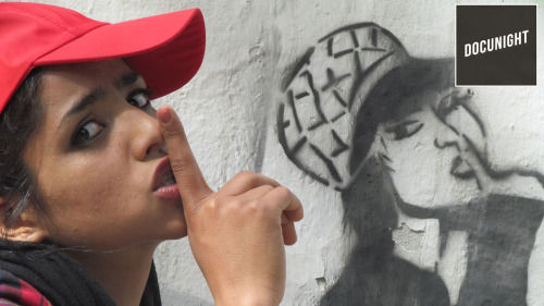 teenage woman wearing a red baseball cap with her index finder covering her mouth to show the sign for quiet